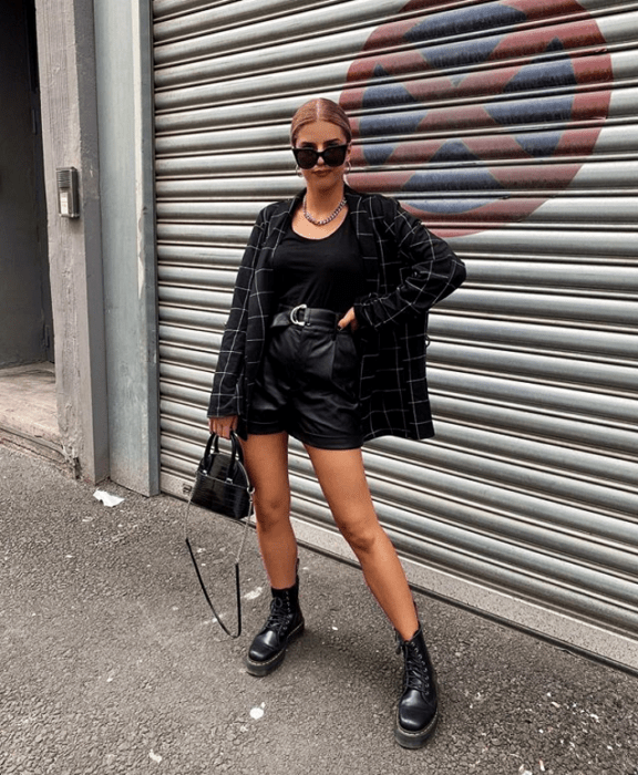chica rubia usando lentes de sol, top negro, blazer saco con líneas blancas, shorts cortos de cuero, bolso pequeño de mano y botines negros de piso