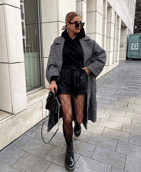 chica rubia usando lentes de sol, sudadera negra, abrigo gris, shorts cortos de cuero negro, medias negras con rombos, bolso de mano negro y botines negros