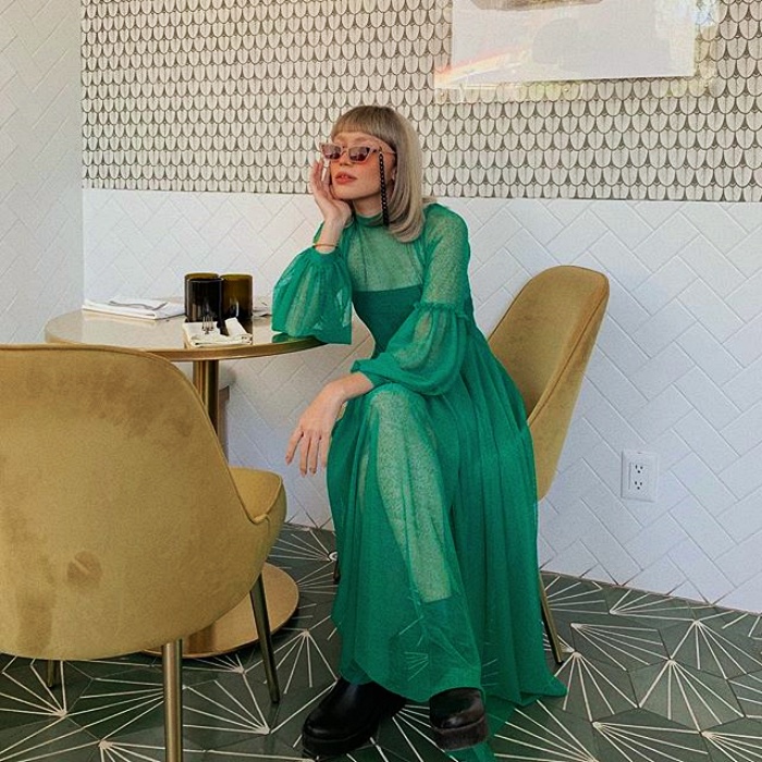 mujer de cabello platinado corto usando gafas de sol rosas, vestido verde con tul de mangas largas y botas negras