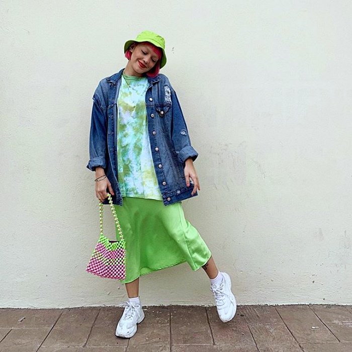 mujer usando un bucket hat verde con chaqueta de mezclilla, camiseta verde con tie-dye y vestido verde de satén, complementado con un bolso rosa y zapatillas deportivas