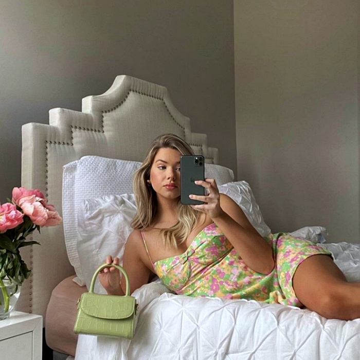 mujer rubia usando un vestido verde pastel con detalles rosas, un bolso mini de mano verde, recostada para tomarse una selfie
