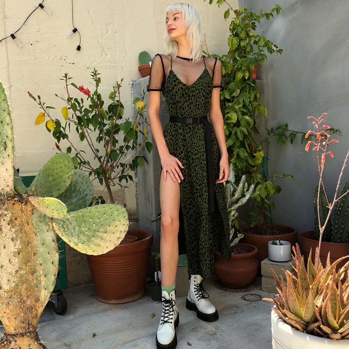 mujer de cabello platinado usando un vestido verde oscuro con tul en las mangas, escote en V y abertura en la pierna, complementado con botas de piel blancas y negras