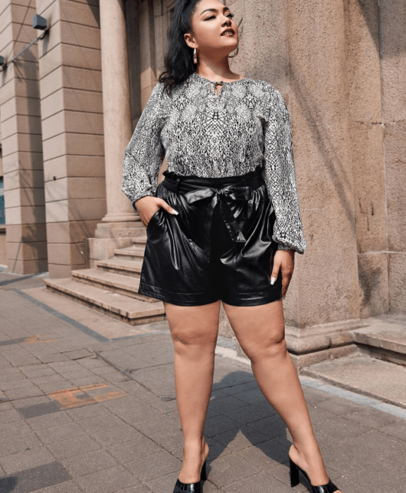 mujer curvy morena con una blusa blanca y negra de manga larga, paperbag shorts negros de cuero y sandalias de tacón negras