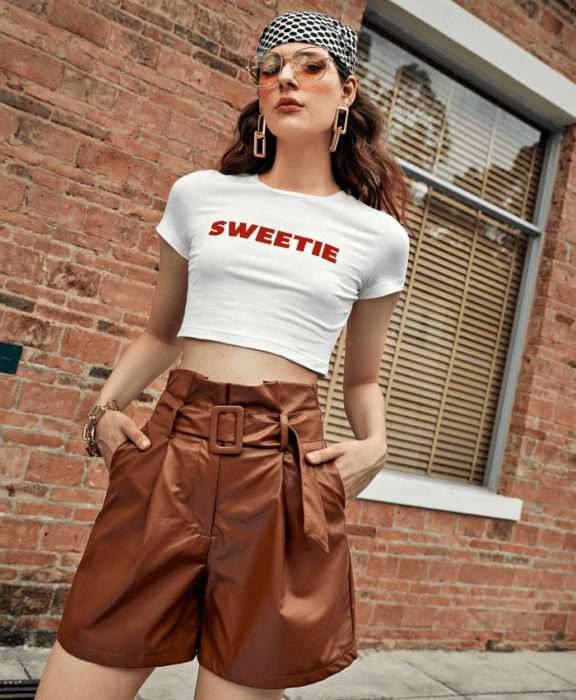 mujer con cabello castaño largo usando gafas de sol y un pañuelo en la cabeza, crop top blanco con letras rojas, paperbag shorts de cuero marrón