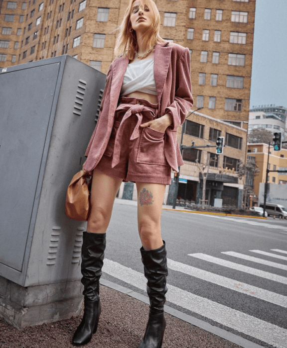 mujer rubia usando un top blanco, saco blazer rosa de pana, paperbag shorts rosas de pana y botas altas de piel y bolso de mano marrón
