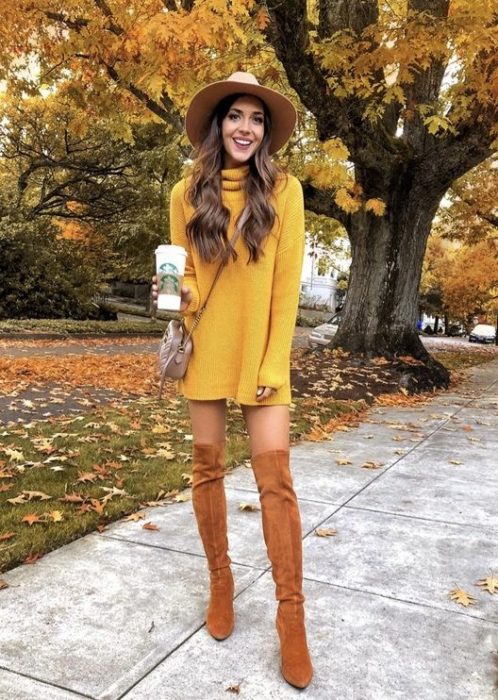 Chica delgada de cabello largo y ondulado con vestido amarillo de manga larga y cuello de tortuga, botas camel y sombrero