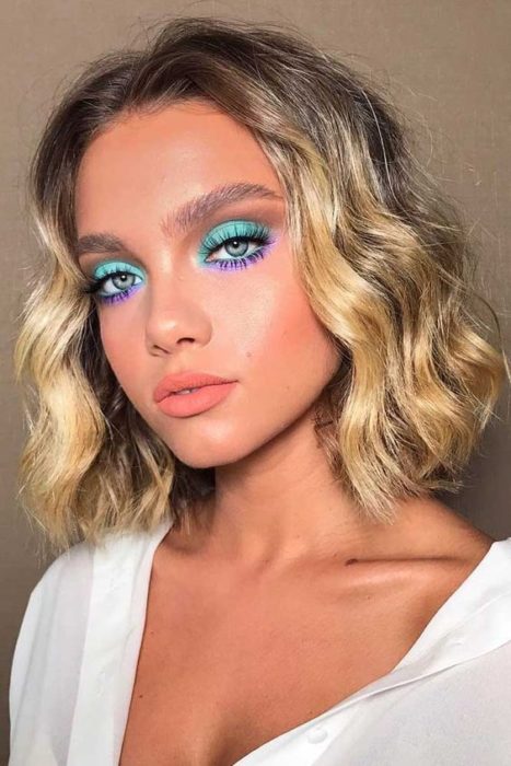Chica rubia de cabello rizado y corto con maquillaje en tonos azul y morado