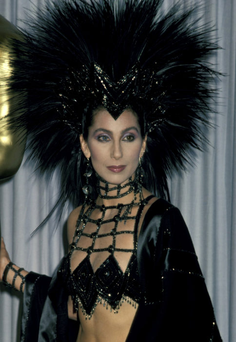 Looks icónicos de Cher en los premios Óscar; tocado de cabello de plumas negras, ropa de red con brillos, ojos de diferente color, azul y verde
