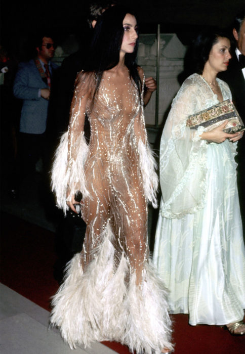 Looks icónicos de Cher; vestido con transparencias, brillos y plumas blancas, cabello lacio, largo y negro