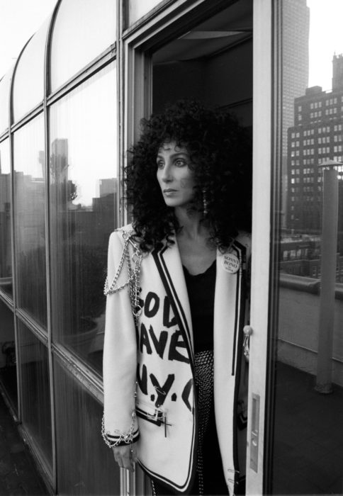 Looks icónicos de Cher; cabello rizado, negro, esponjado, saco blanco moderno con cadenas y frase