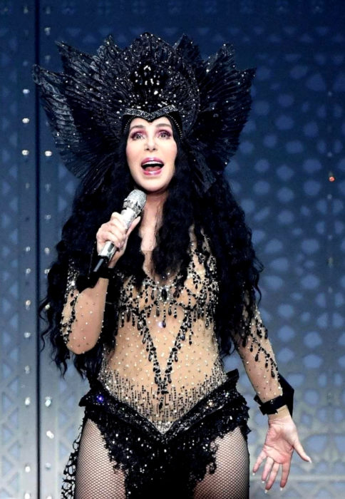Looks icónicos de Cher; cabello negro, largo, rizado, tocado de plumas y alas, en concierto