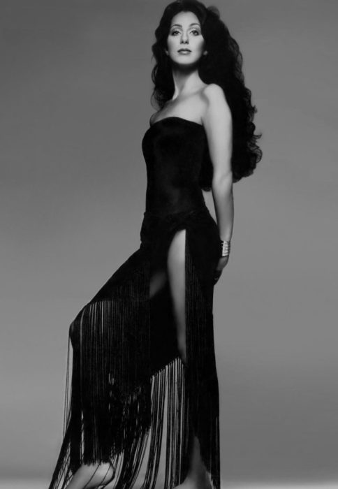Looks icónicos de Cher; cabello negro, rizado, largo, vestido negro de cocktail