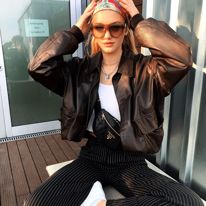 chica rubia usando lentes de sol, pañoleta en el cabello, chamarra de cuero café, top blanco, cangurera de cuero, pantalones de vestir azules con lineas blancas