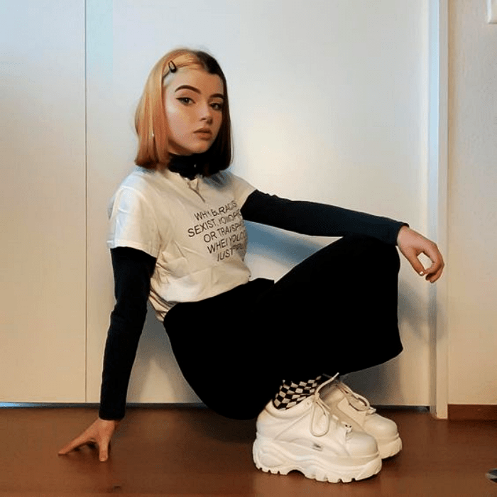 chica con cabello bicolor negro y rubio usando una camiseta blanca con mangas negras y pantalón acampanado negro, tenis deportivos con plataforma blancos