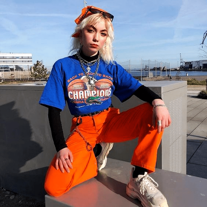 chica rubia usando lentes de sol rojos, camiseta azul con estampado, mangas negras, pantalón acampanado anaranjado y tenis deportivos blancos
