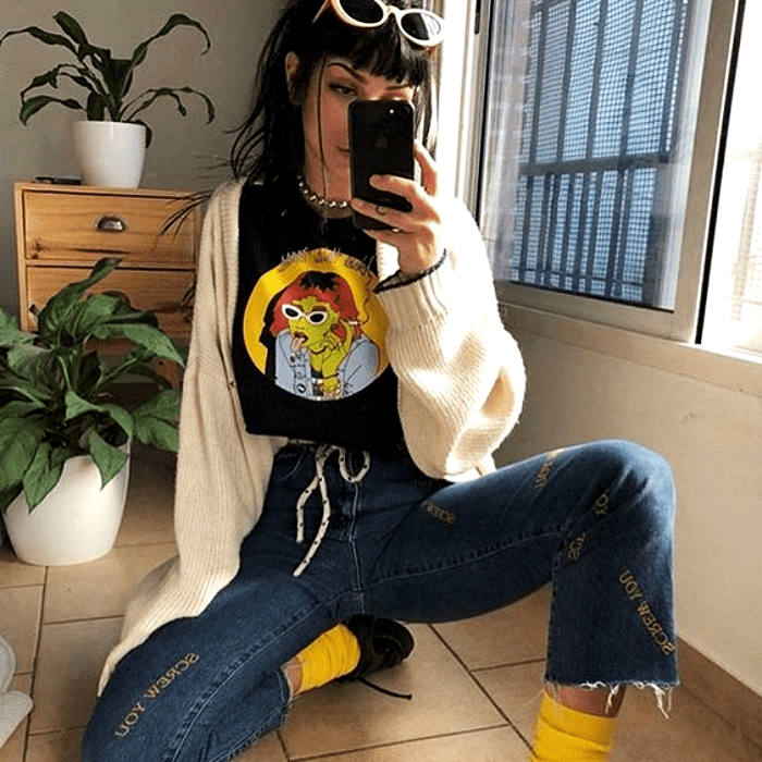 chica con cabello oscuro usando lentes de sol blancos, camiseta negra con estampado, suéter cárdigan blanco, pantalón acampanado de mezclilla, calcetas amarillas y zapatos de charol con plataforma