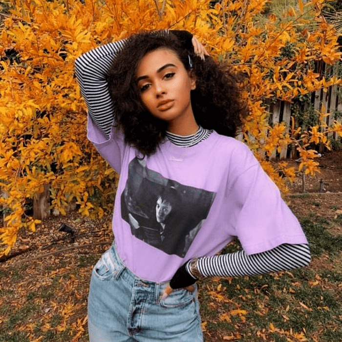 chica morena con cabello chino, usando una camiseta lila con mangas grises, pantalon de mezclilla a la cintura