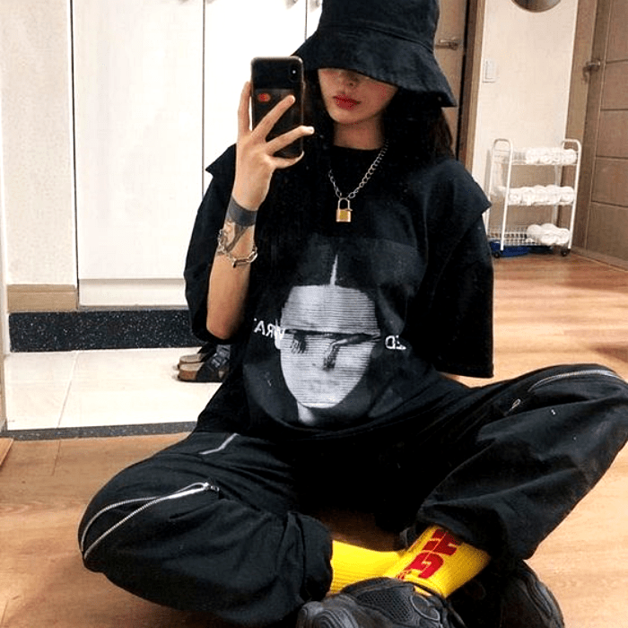 chica de cabello negro largo usando un bucket-hat negro, camiseta negra con estampado gris, pantalones baggy negros, calcetas amarillas largas y tenis deportivos negros