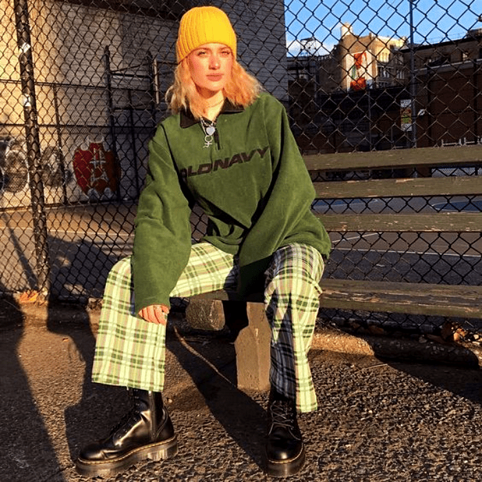chica con cabello rosa usando una gorra amarilla, sudadera verde y pantalones blancos con cuadros verdes, botas de cuero de plataforma