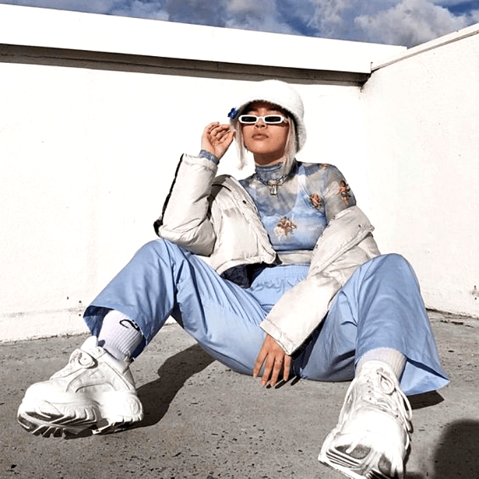 chica con cabello platinado usando un bucket-hat blanco, top con estampado de nubes, chamarra blanca, pantalones azul celeste baggy y tenis deportivos blancos