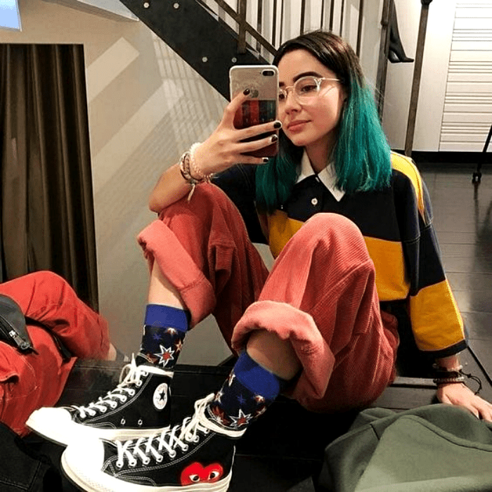 chica con cabello teñido de color azul usando lentes, camiseta de manga larga con rayas amarillas y negras, pantalon rosa, converse negros de botin
