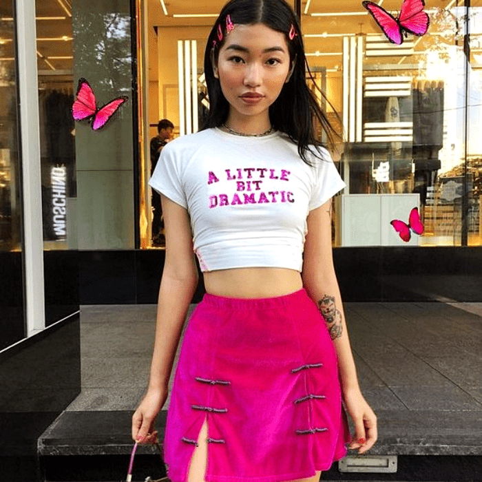 chica con cabello castaño largo usando un crop-top blanco con letras rosas, falda rosa brillante y bolso de mano