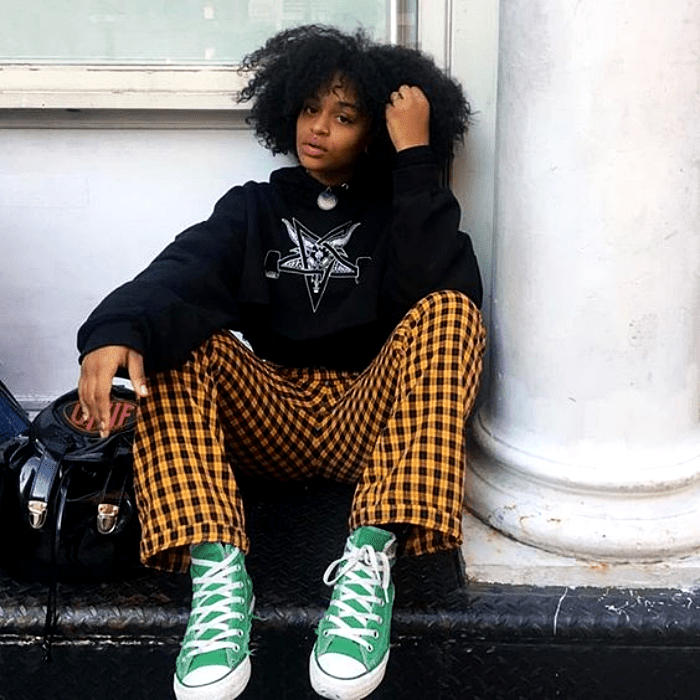 chica morena con cabello chino usando una sudadera negra con estampado y pantalones amarillos con cuadros negros, converse verdes