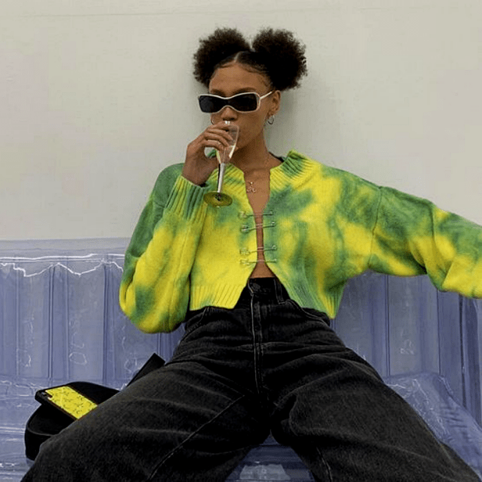 chica morena con cabello castaño usando un suéter de tie dye verde con amarillo, pantalón verde a la cintura y lentes de sol