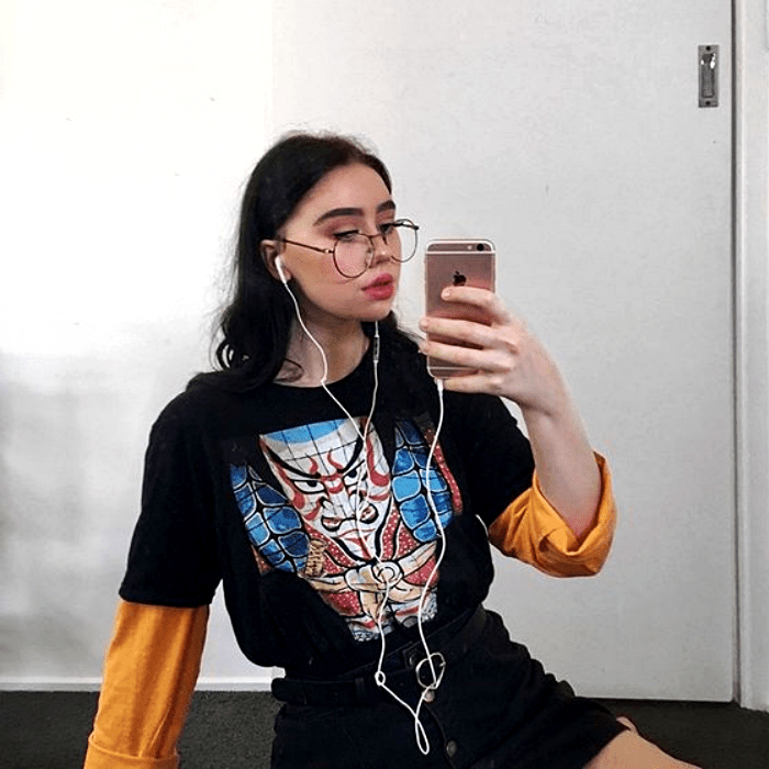 chica de cabello castaño con lentes usando una camiseta negra con estampado, mangas anaranjadas y minifalda negra