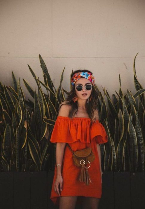 Chica usando outfit veraniego de vestido naranja off the shoulder, arriba de la rodilla, con lentes oscuros, banda en el cabello y bolso cruzado