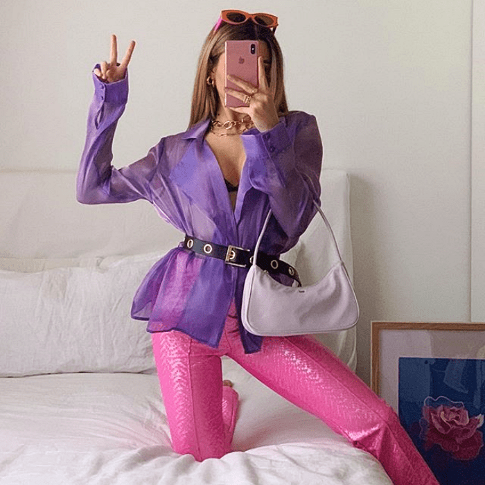 chica rubia usando blusa de gaza morada con rosa, bolso de mano lila, pantalones de cuero rosas