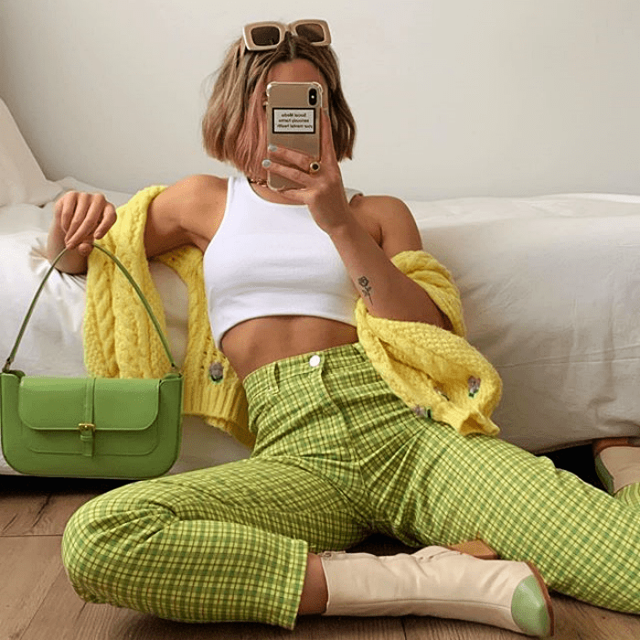 chica rubia usando top blanco crop-top, suéter amarillo, pantalones verdes de cuadros, botines beige con punta verde, bolso de mano pequeño verde