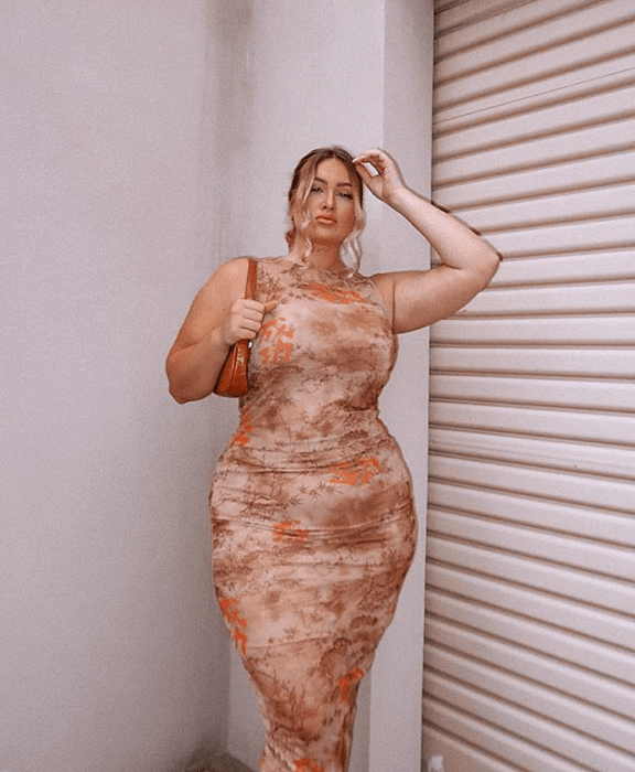 Chica rubia curvy en un vestido ceñido color nude con estampado de rosas y flores, usando un bolso de mano naranja