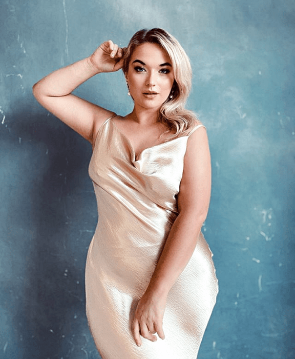 Chica rubia platinada curvy en un vestido color champán ceñido de satén con tirantes y escote holgado