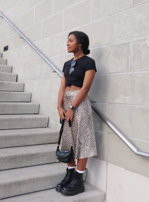 Chica morena con blusa negra y falda de estampado de leopardo sosteniendo un bolso negro y usando botas dr. martens