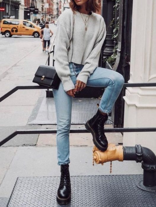 Chica sentada en un barandal con suéter gris oversized, jeans azules, bolso negro cuadrado en el hombro y botas dr. martens negras