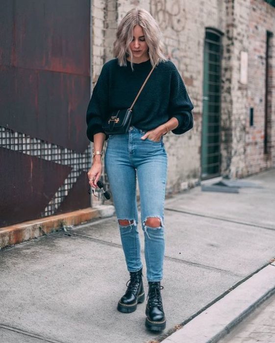 Chica rubia de melena corta con suéter azul marino oversized y jeans rasgados de las rodillas con botas dr. martens negras
