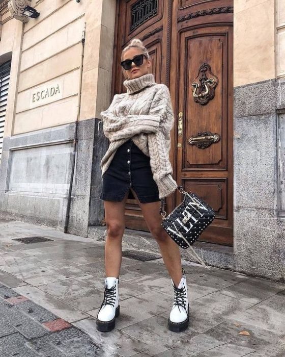 Chica rubia de cabello recogido en un chongo con lentes de sol negros y suéter oversized beige, falda negra con botones en la parte delantera y botas dr. martens blancas