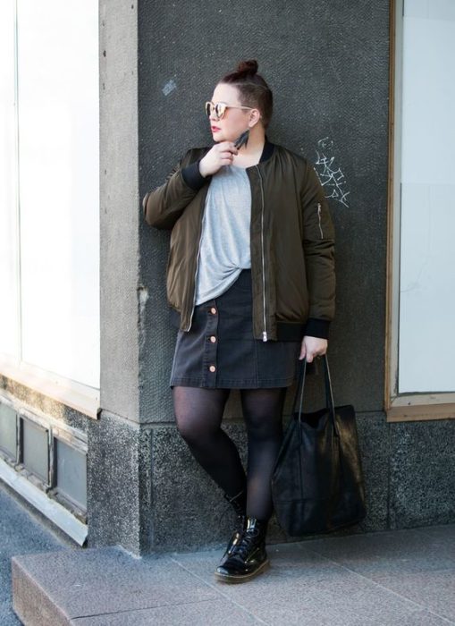Chica plus size recargada en la pared con chaqueta verde militar, blusa gris, falda negra, medias negras y botas dr. martens