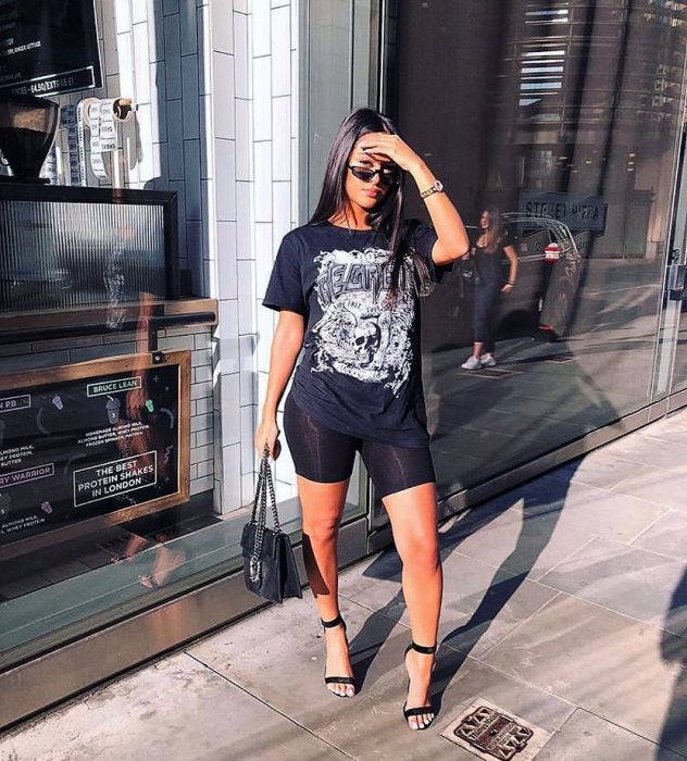Chica morena de cabello largo y suelto se tapa el sol con la mano y usa blusa y shorts de ciclista color negros