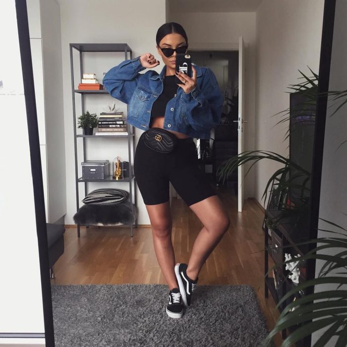 Chica con el cabello recogido en un chongo se toma selfie frente al espejo con chamarra de mezclilla y shorts de ciclista negros