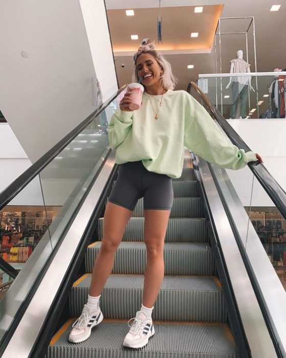 Chica rubia en escaleras eléctricas sosteniendo un café mientras usa sudadera verde pistache y shorts de ciclista grises con tenis blancos
