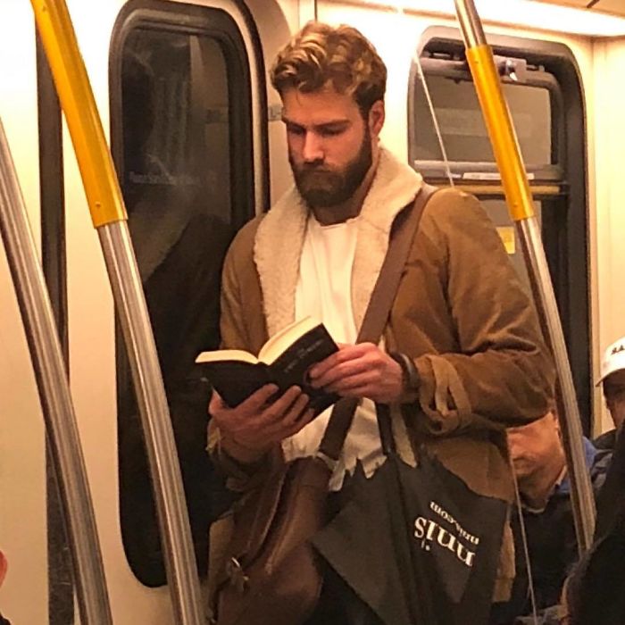 Chico con barba abultada recargado en la puerta del metro leyendo un libro