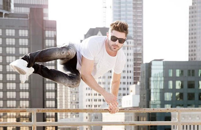 Zac Efron sosteniéndose con las manos en un barandilla