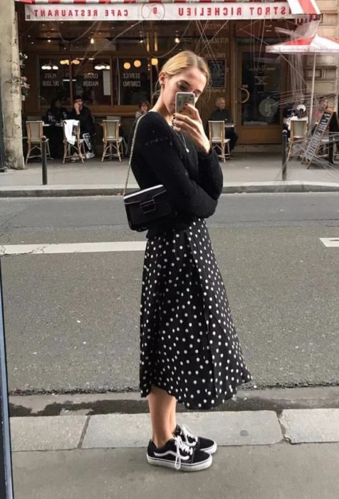Chica rubia tomando una selfie con suéter y falda negros con tenis negros vans
