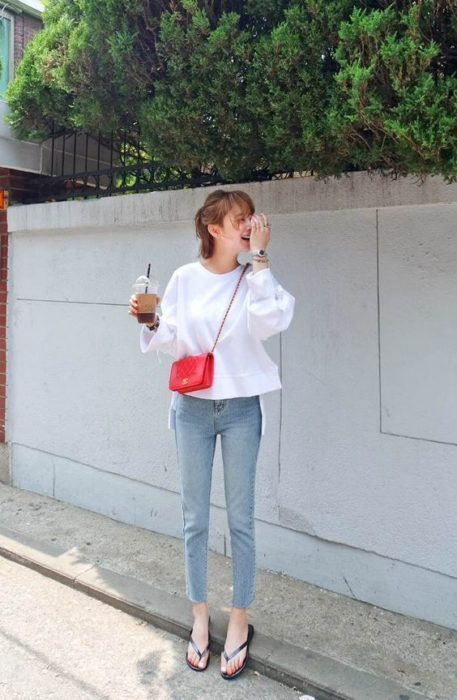 Chica de cabello corto sostiene una coca cola y usa jeans, sudadera blanca y flip flops