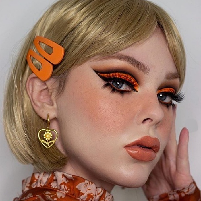 chica rubia con maquillaje sesentero, sombra de ojos color naranja, delineado cat-eye negro, lipstick naranja
