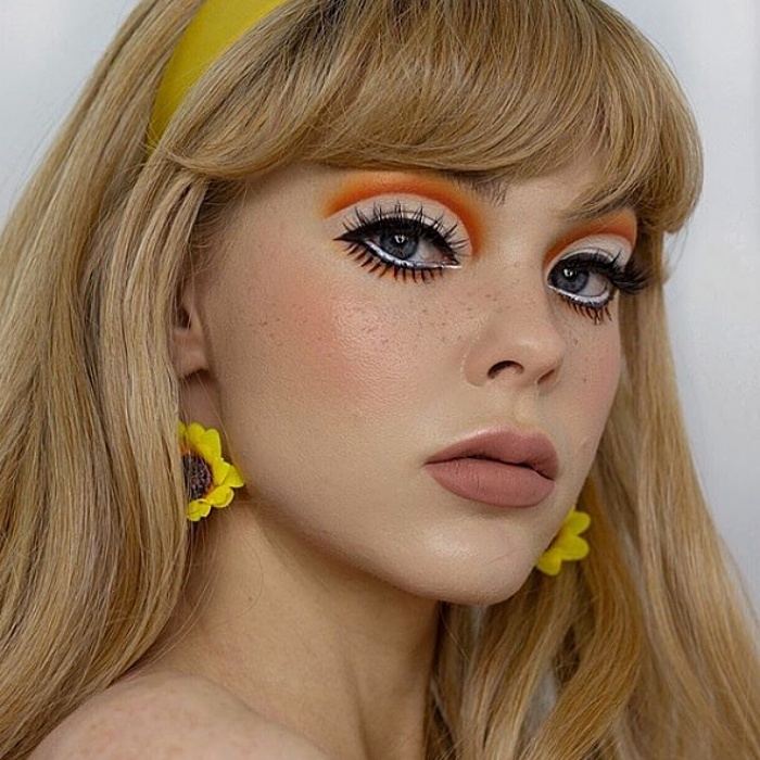 chica con cabello rubio largo, usando maquillaje tipo de los sesentas, delineado con color naranja, pestañas postizas, aretes de girasol