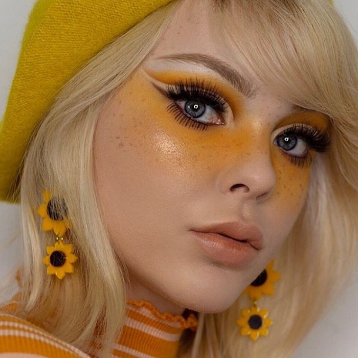 chica rubia usando maquillaje con sombras amarillas, delineado negro oscuro y labios naturales, usando boina amarilla y aretes de girasol