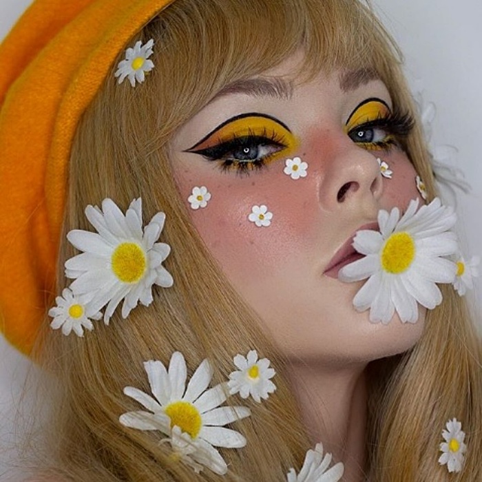 chica rubia de cabello largo usando margaritas flores en el cabello, con maquillaje de sombra de ojos amarilla, delineado negro, blush rosa y boina anaranjada